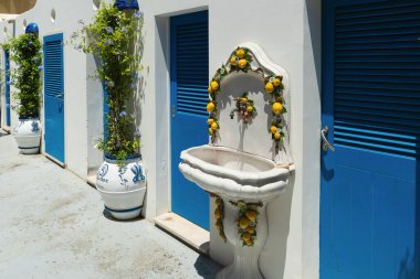 Positano, İtalya - 15 Haziran 2024: Limon temalı bir çeşme, güneşli bir günde Amalfi Sahili 'ndeki pastel binalara cazibe katıyor. Canlı mavi kepenkler bu resim görünümünü tamamlıyor.