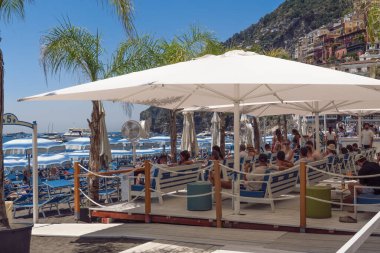 Positano, İtalya - 15 Haziran 2024: Ziyaretçiler güneşli bir gökyüzünün altındaki Amalfi Kıyısı 'nın berrak mavi sularının kıyısındaki bir plaj kafesinde dinleniyorlar..