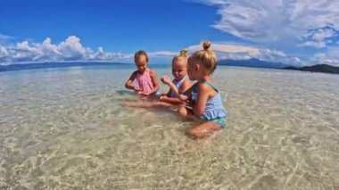 Üç küçük kız 4 yaşında, koşuyor ve mayoyla oynuyor okyanusta bir kum tepesinde, sığ su, temiz su, sarı kum, bulutlar Triplet 'in çocukları sahilde oynuyorlar 