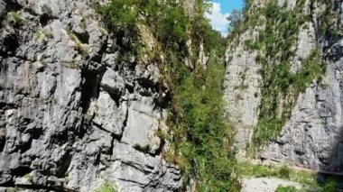 Yazın dar bir dağ geçidi, bir dron geçitten uçuyor, duvarlar yüksek ve taştan, yeşil ağaçlar yetişiyor, gökyüzü bulutsuz, güneş parlak. Yüksek kalite 4k görüntü