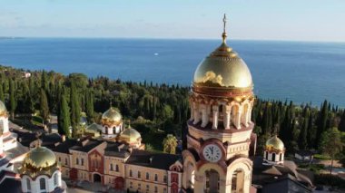 Abhazya 'daki yeni Athos Manastırı, Yeni Athos şehri, İHA görüntüleri, sonbahar yeşil tepeleri ve mavi deniz, batan güneşte parıldayan kiliselerin altın kubbeleri, Ortodoks haçları, selvi ağaçları ve palmiye ağaçları.