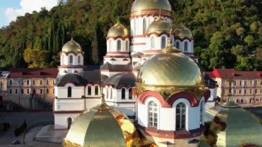 Abhazya 'daki yeni Athos Manastırı, Yeni Athos şehri, İHA görüntüleri, sonbahar yeşil tepeleri ve mavi deniz, batan güneşte parıldayan kiliselerin altın kubbeleri, Ortodoks haçları, selvi ağaçları ve palmiye ağaçları.