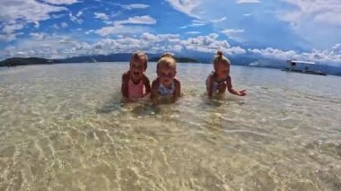 Üç küçük kız 4 yaşında, koşuyor ve mayoyla oynuyor okyanusta bir kum tepesinde, sığ su, temiz su, sarı kum, bulutlar Triplet 'in çocukları sahilde oynuyorlar 