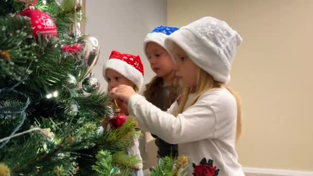 三胞胎的三个妹妹用玩具和花环装饰圣诞树 孩子们戴着五颜六色的圣诞老人帽 圣诞树旁边有一个家庭壁炉在燃烧 — 图库视频影像