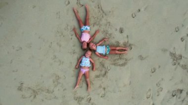 Üç kız kardeş sırtüstü gökyüzüne bakıyor, 3 yaşında küçük kızlar, üçüzler, beyaz kum ve okyanus, mayo üçüzleri kumun üzerinde uzanıp gökyüzüne bakıyor. Yüksek kalite 4k dron