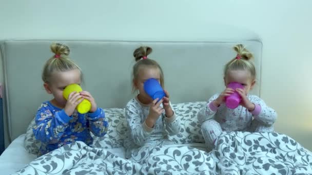 Три Маленькие Сестры Тройняшки Пьют Молоко Разноцветных Стаканов Сидя Постели — стоковое видео