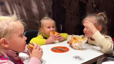 Bir fast food restoranında elleriyle hamburger yiyen ikiz üçlü kız kardeşler, kalın kazaklar ve kayışlı pantolonlar giyiyorlar. Yüksek kalite 4k görüntü