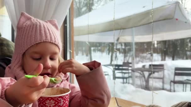窓の外にココアのプラスチックガラスから緑色のスプーンでマシュマロを食べる小さな女の子は雪の冬です 高品質の4K映像 — ストック動画