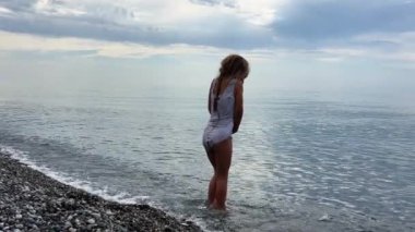 Küçük kız denizin soğuk sularında çakıl taşı plajında duruyor, bulutlu gökyüzü sakin. Yüksek kalite 4k görüntü