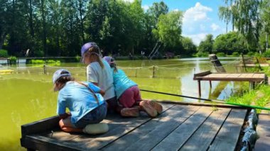 Gölün rıhtımında balık tutmak için oturan üç küçük kız kardeş sudaki balık oltalarına bakarlar. Yüksek kalite 4k görüntü