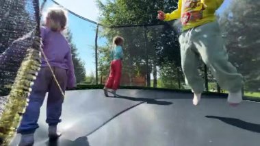 Üç küçük kız kardeş üçüzler yazın sokakta trambolinde zıplıyorlar kızlar 4 yaşında, kazak ve pantolon giyiyorlar, ağaçların arkasındaki trambolinde bir ağ gerilmiş.