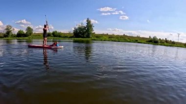 Huzurlu bir göl sahnesinde, sakin suda zarif bir şekilde kürek çeken bir kadın, ardından sükunetin tadını çıkaran, güzelliğini sergileyen bir erkek görülüyor.