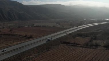 Türkiye 'de otoyol boyunca beyaz bir karavan sürüyor, İHA görüntüleri, kurumuş tarlalar ve kahverengi dağlar, güneş parlıyor. Yüksek kalite 4k görüntü