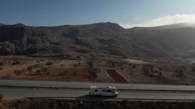 Türkiye 'de otoyol boyunca beyaz bir karavan sürüyor, İHA görüntüleri, kurumuş tarlalar ve kahverengi dağlar, güneş parlıyor. Yüksek kalite 4k görüntü