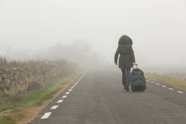 Sisli bir yolda yürüyen, bavulu çeken biri gizemli ve macera dolu bir atmosfer yaratır..