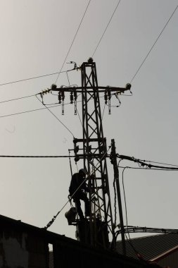 Siluetli bir işçi günbatımında gerekli elektrik bakımını yapmak için ustalıkla uzun bir elektrik direğine tırmanır.
