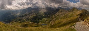 Arka planda bulutlu bir gökyüzü olan bir dağ sırası. Dağlar çimlerle kaplı ve ön planda bir göl var.