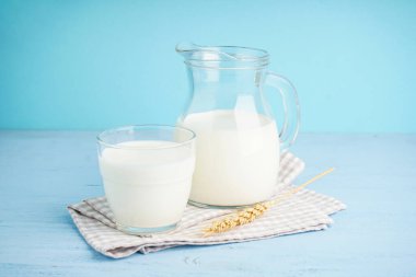 Açık mavi arka planda sürahi ve bir bardak taze süt ve mutfak havlusu..