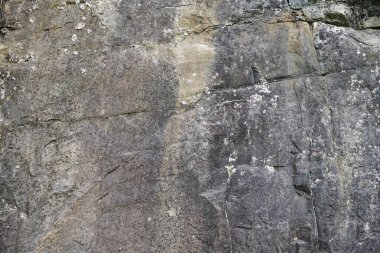 Doğal kaya yüzeyinin çatlaklardan, katmanlardan, yosun ve liken parçalarından oluşan dokusu ile yakın çekim. Gri-kahverengi tonlardaki doğal taş arka plan olarak idealdir..