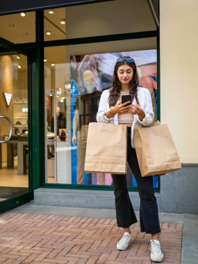 Alışveriş merkezinin önünde cep telefonuna bakan genç beyaz bir kadın. Kollarından sarkan çantalar. İndirim gününde alışveriş. Kara Cuma. Malaga 'daki Plaza Belediye Başkanı alışveriş merkezi