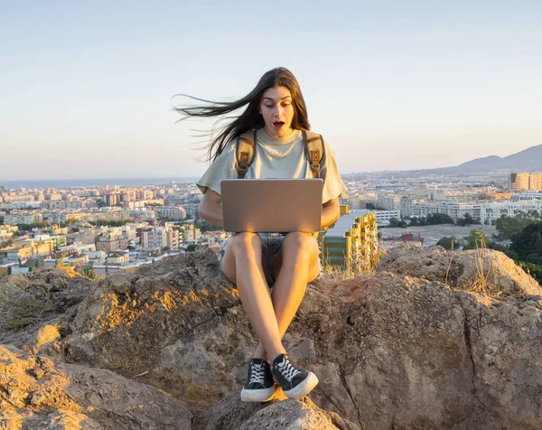 Young woman with a backpack, using a laptop to make a video call in the middle of a field. Digital nomad. In the background Malaga at sunset.