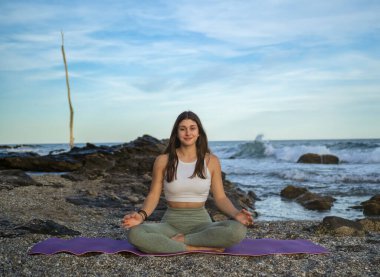 Nilüfer pozisyonunda oturan ve Akdeniz sahilinde sağlıklı bir yaşam tarzı için yoga yapan genç bir kadın. Kadın gücünü ve ruhunu geri kazanmak için spor egzersizleri yapıyor. Yoga pozisyonu gün batımında deniz kıyısında.