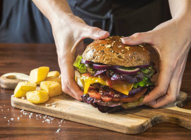 Koyu ahşap bir masada sebzeli ve patates kızartmalı leziz bir hamburgerin yakın plan çekimi. Tanınmayan biri hamburgeri iki eliyle alır ve yer..