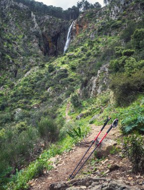 Akan bir şelaleye giden dağ yolu. Ön planda kırmızı bastonlar. Yeşil bitki ve çalılarla dolu sakin ve serin bir ortam..