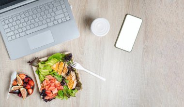 Bilgisayarlı çalışma alanı, sağlıklı yemek servisi, ormanın meyvesi ve somon ve avokadoyla salata. Tampondaki yiyecekleri al. Yukarıdan görüntüle, arkaplanda tahta masa.