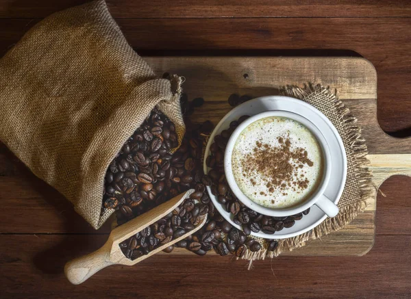 stock image Top view of a cup of coffee on a wooden board and a sack full of coffee beans. A spoon with coffee beans. Warm colors. Copy space.