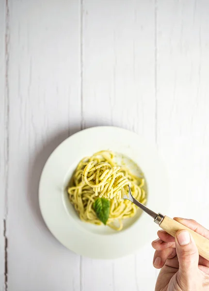 Taze pesto soslu spagetti italiani. Spagettiyle çatal tutan bir el. Hepsi beyaz ahşap bir masada..