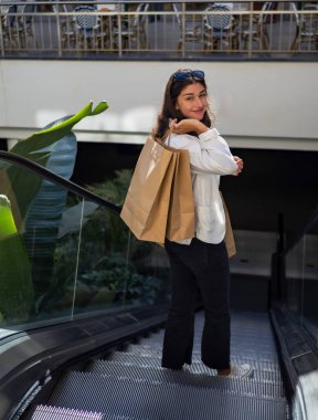 Alışveriş merkezinden alışveriş yaptıktan sonra sırtında alışveriş torbalarıyla yürüyen merdivenden inen genç bir kadın. Kara Cuma, kelepir.