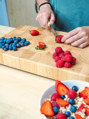Mutfaktaki kadın, tahtadan bir masada mısır gevreği ve böğürtlenle sağlıklı bir atıştırmalık hazırlamak için meyve kesiyor..