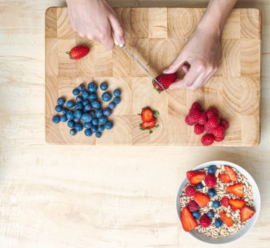 Mutfaktaki kadın, tahtadan bir masada mısır gevreği ve böğürtlenle sağlıklı bir atıştırmalık hazırlamak için meyve kesiyor..