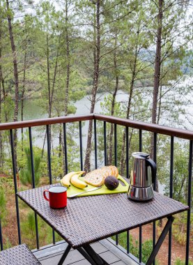 Avokado, muz, ekmek ve kahveyle vejetaryen kahvaltısı, arka planda bir orman ve gölle birlikte terasta yenecek. Sağlıklı bir yaşam tarzı.