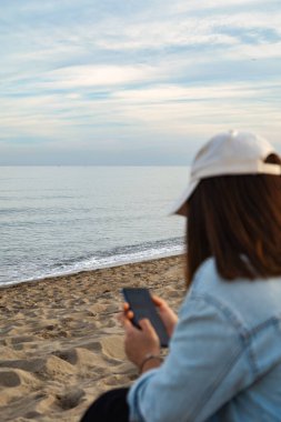 Odaklanamayan kız deniz kıyısındaki sahilde oturan cep telefonuna bakıyor..