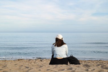 Girl with cap sitting on the seashore of a beach without people, feeling of serenity and solitude. clipart