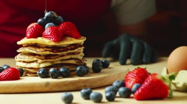 Önlüklü ve siyah eldivenli bir adam kremalı, çilekli ve böğürtlenli kreplerin üzerine bal döküyor.