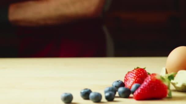 戴着围裙和黑色手套的男人 在木板上摆放着奶油 草莓和蓝莓煎饼 有着温暖的光线和横向的视野 — 图库视频影像