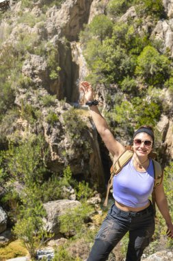 Kameralı sırt çantasına bakan kız koluyla vadiden, turizmden, maceradan ve doğadan bir şelaleyi işaret ediyor.