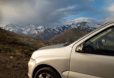 Araba şafak vakti yüksek bir dağ yolunda otlayan koyunlar ve arka planda karla kaplı zirvelerle park edilmiş.