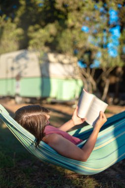 Hamakta yatan bir kadın, bir karavanın arka planında kitap okuyor, kırsal huzur..