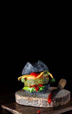 Siyah ekmekli hamburger, çedar peyniri, domates ve tahtanın tepesinde ıspanaklı et ve siyah arka planda ketçaplı bir bıçak, cadılar bayramı tarzı..