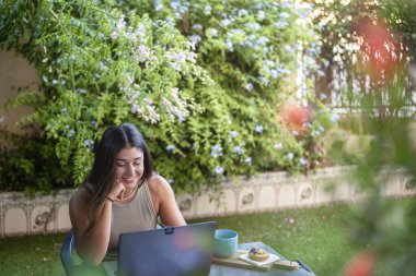Gülümseyen kız evinin bahçesinde dizüstü bilgisayarın önünde oturuyor yanında atıştırmalık bir şeyler var..