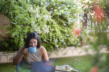 Dizüstü bilgisayarın önünde içki içen bir kız kupa ve telgrafla evinin arka bahçesinde çalışıyor..