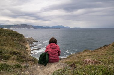 Kırmızı ceketli beyaz bir kadın sessiz bir kayanın üzerinde oturuyor ve kayalıklarda ve deniz dalgalarında dinleniyor, Bask Ülkesi, İspanya