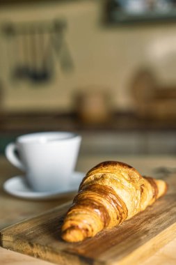 Kırsal mutfak masasında ayçöreği olan bir fincan kahve. Tahta üzerinde ön planda taze kruvasanlar. Evde kahvaltı, sıcak ışık ve rahat atmosfer..
