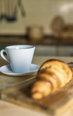 Kırsal mutfak masasında ayçöreği olan bir fincan kahve. Ön planda tahtadan yapılmış taze kruvasanlar. Evde kahvaltı, sıcak ışık ve rahat atmosfer..
