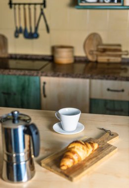 Mutfakta kahvaltı, kahve demliği ve sıcak kahve, boş bardak ve kruvasan tahtada, arka planda mutfak. Sıcak ışık ve sıcak atmosfer