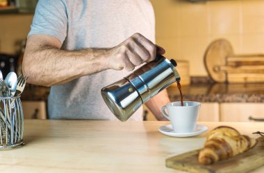 Yeni uyanan beyaz adam bir İtalyan kahve makinesiyle kahve servisi, kruvasanla kahvaltı. Mutfakta sıcak ışık ve sıcak atmosfer var..